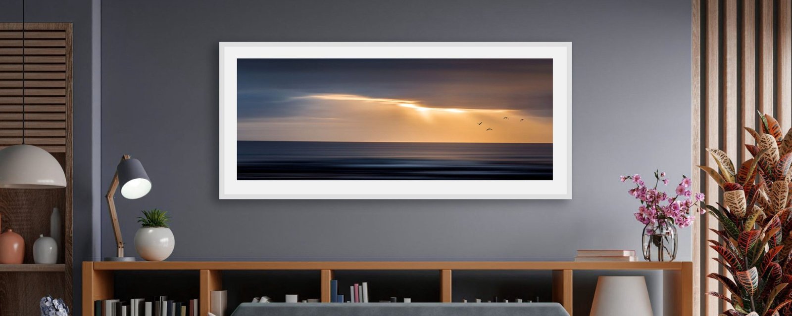 A sunset seascape in a wide white frame above a low bookcase